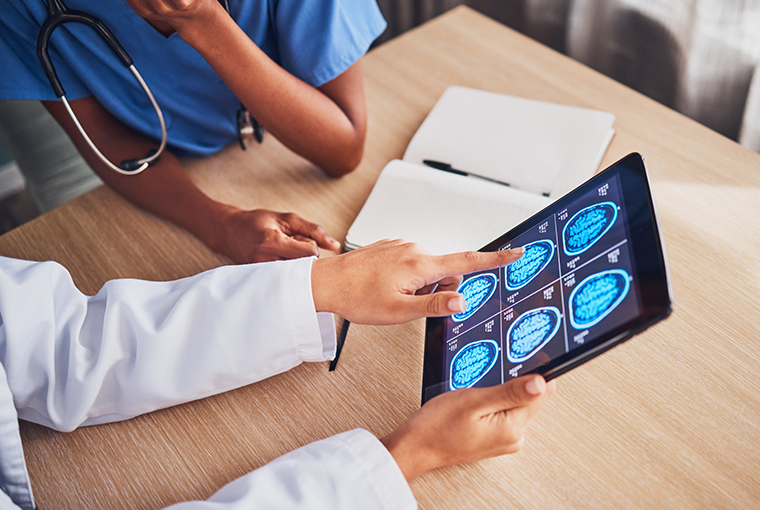 Ein Tablet zeigt Gehirnscans, während zwei Personen – eine in weißem Kittel und eine in blauer Kleidung – die Bilder an einem Schreibtisch mit Notizbuch besprechen, wobei sie die möglichen neurologischen Störungen durch Vitamin-B12-Mangel thematisieren.
