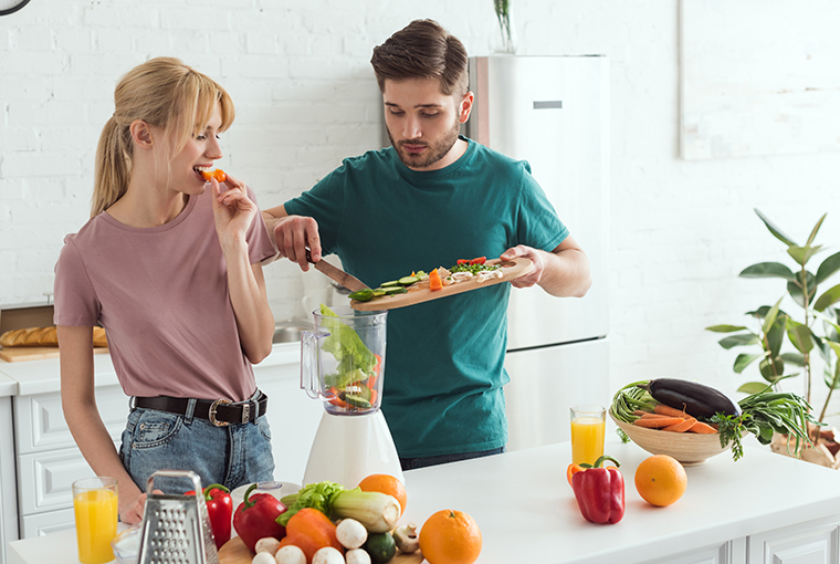 Ein Mann fügt Gemüse in einen Mixer ein, während eine Frau in der hellen Küche, umgeben von Obst und Gemüse, daran knabbert; Vitamin B12 ist besonders wichtig bei pflanzlicher Ernährung.