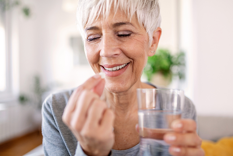 Eine Frau hält in einer Hand eine Vitamin-B12-Tablette und in der anderen ein Wasserglas, während sie in ihrem sonnendurchfluteten Wohnzimmer mit Pflanzen im Hintergrund steht.