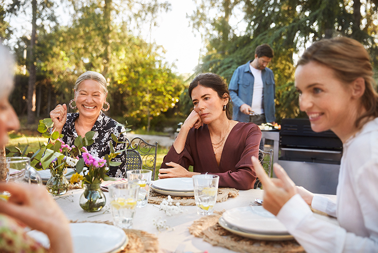 Eine Frau mit einer gestörten Aufnahme von Vitamin B12 sitzt antriebslos an einem Gartentisch mit zwei anderen Menschen.
