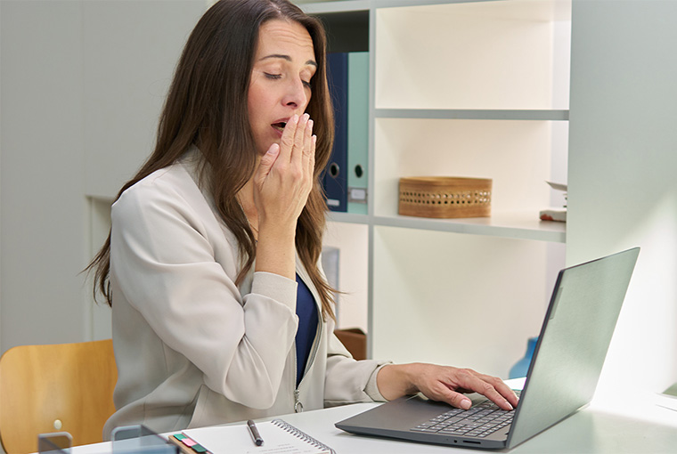 Eine müde Frau sitzt an einem Bürotisch mit zwei anderen Personen und zeigt typische Symptome eines Vitamin B12 Mangels.